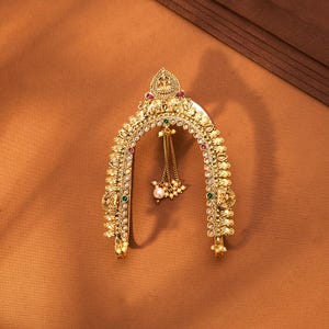 May include: Ornate gold-toned headpiece with intricate detailing, featuring embedded red, green, and white gemstones. The arched design includes dangling chains with pearls and small beads. The headpiece is displayed on a brown surface.