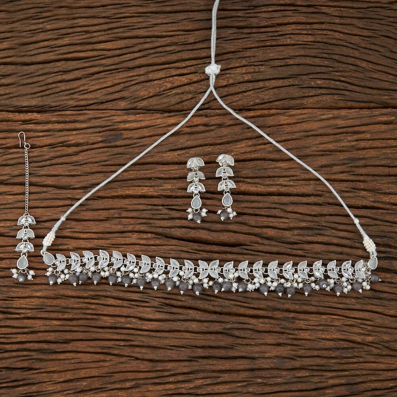 May include: A silver-toned jewelry set featuring a necklace, earrings, and a headpiece. The necklace and headpiece have leaf-shaped accents and gray beads. The earrings have a similar design. The set is displayed on a wooden surface.