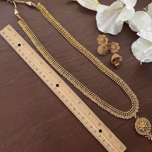 May include: A gold-toned necklace with coin-shaped pendants and a matching pair of earrings. The necklace, approximately 28 inches long, features a detailed pendant at the bottom. The earrings have a floral design. A ruler is placed next to the jewelry.