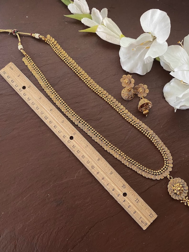 May include: A gold-toned necklace with coin-shaped pendants and a matching pair of earrings. The necklace, approximately 28 inches long, features a detailed pendant at the bottom. The earrings have a floral design. A ruler is placed next to the jewelry.