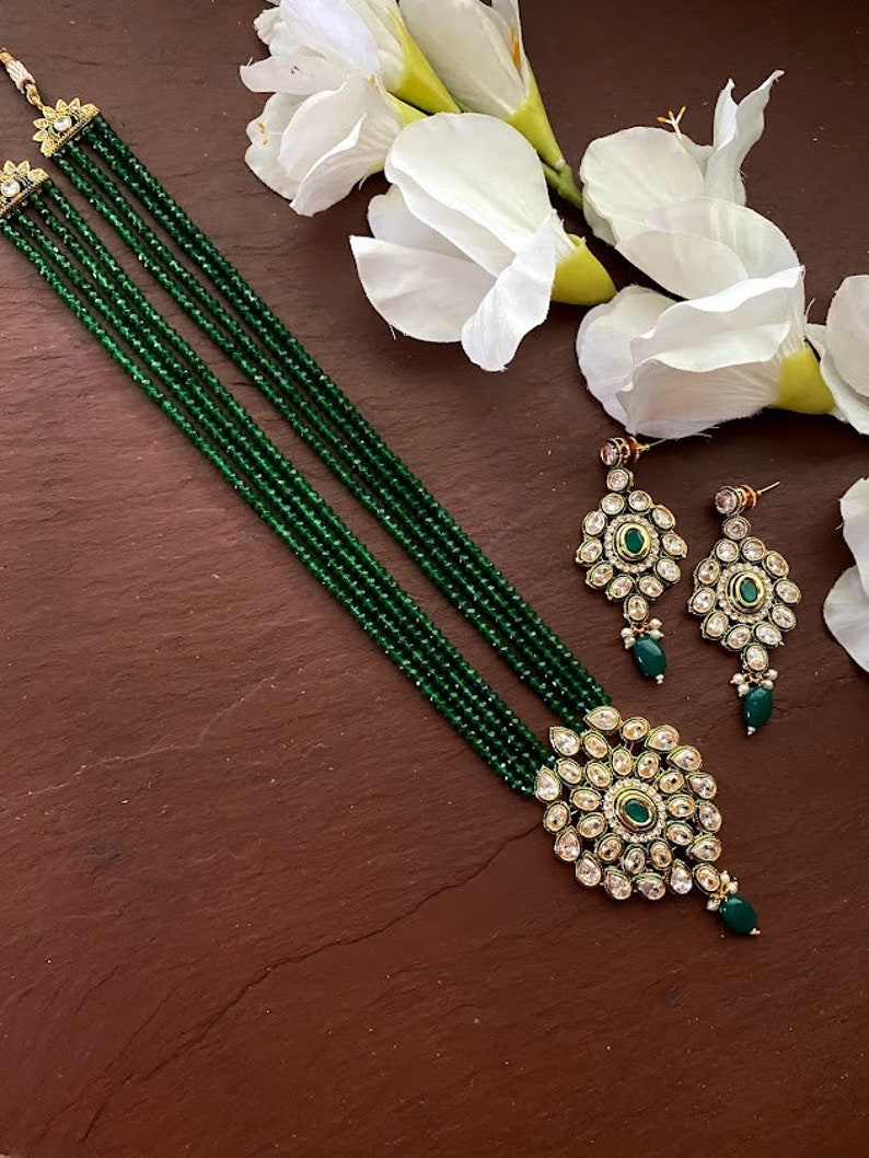 May include: A green beaded necklace with a large, ornate pendant featuring a green stone surrounded by clear stones. The pendant is attached to a gold chain. Matching earrings with a similar design are also included.