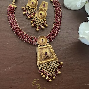 May include: A gold necklace with a red bead chain and a pendant with a square design. The pendant has a small red stone in the center. The necklace comes with matching earrings with a round design and a red stone in the center.