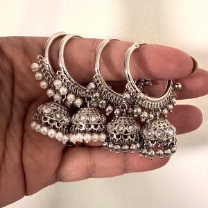 May include: Four silver-toned hoop earrings with intricate detailing. Each earring features a bell-shaped charm adorned with clear stones and small white pearls. The earrings are held in a hand, showcasing their design.