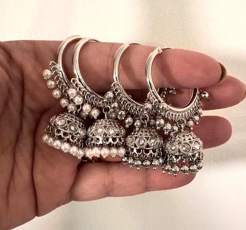 May include: Four silver-toned hoop earrings with intricate detailing. Each earring features a bell-shaped charm adorned with clear stones and small white pearls. The earrings are held in a hand, showcasing their design.