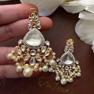May include: A pair of gold earrings with a teardrop-shaped design. The earrings are adorned with clear stones and pearls.