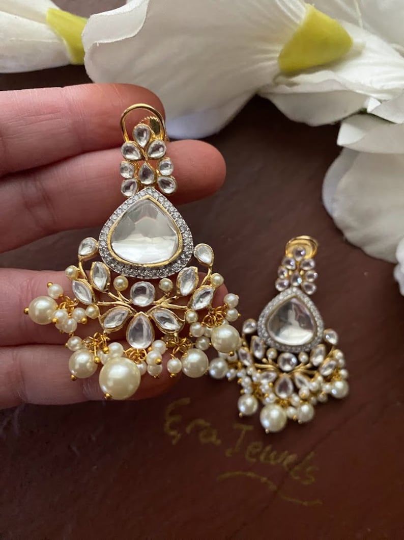 May include: A pair of gold earrings with a teardrop-shaped design. The earrings are adorned with clear stones and pearls.