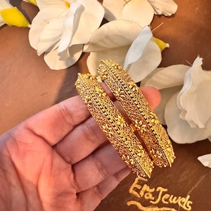 May include: Two gold-colored bangles with intricate floral and geometric patterns. The bangles are held in a hand, with white flowers in the background. The text "Era Jewels" is visible.