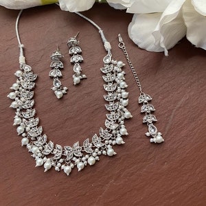 May include: A silver-toned jewelry set featuring a necklace, earrings, and a headpiece. The set includes leaf-shaped accents with clear stones and dangling faux pearls. The necklace and headpiece are connected by a delicate chain.