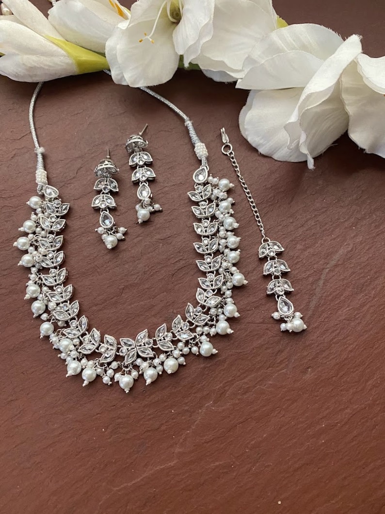 May include: A silver-toned jewelry set featuring a necklace, earrings, and a headpiece. The set includes leaf-shaped accents with clear stones and dangling faux pearls. The necklace and headpiece are connected by a delicate chain.
