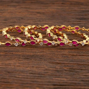 Puede incluir: Conjunto de cuatro brazaletes de oro con piedras rosas y blancas. Los brazaletes están apilados uno encima del otro y están sobre una superficie de madera.