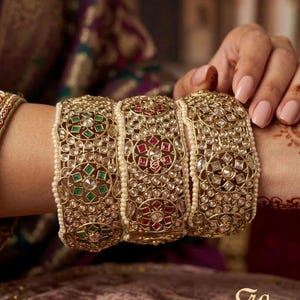 May include: Gold-toned bangles with intricate designs, featuring embedded gemstones in green and red, and accented with small pearls. The bangles are worn on a wrist, showcasing the detailed craftsmanship and ornate style.