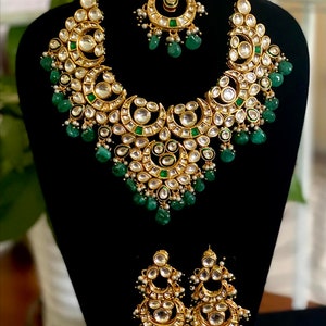 May include: A gold-toned jewelry set featuring a necklace, earrings, and a headpiece. The necklace and earrings have a crescent moon design with clear and green stones. The set is displayed on a black mannequin.