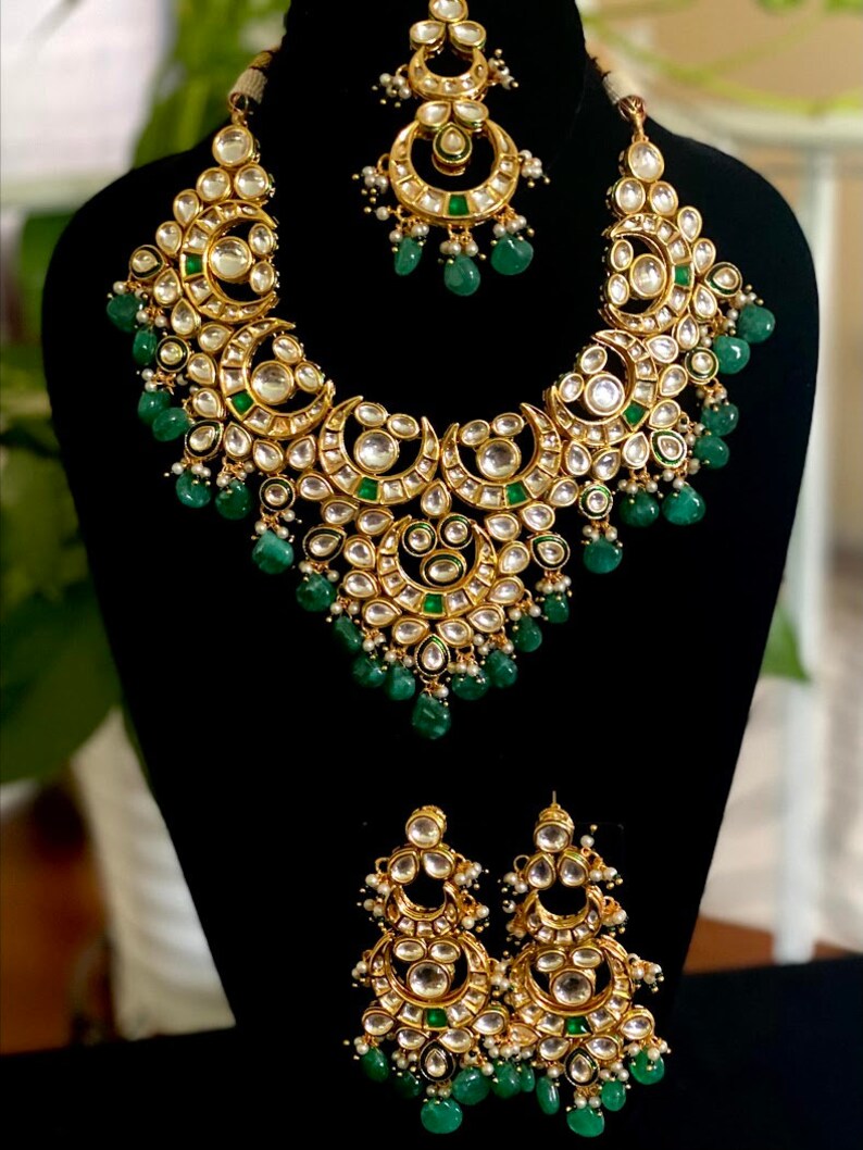 May include: A gold-toned jewelry set featuring a necklace, earrings, and a headpiece. The necklace and earrings have a crescent moon design with clear and green stones. The set is displayed on a black mannequin.