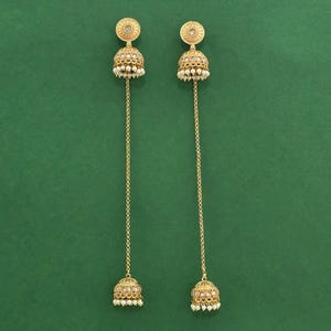 May include: A pair of gold-colored earrings with a floral design and small white pearls. The earrings have a long chain that connects to a bell-shaped charm at the bottom. The background is a solid green color.