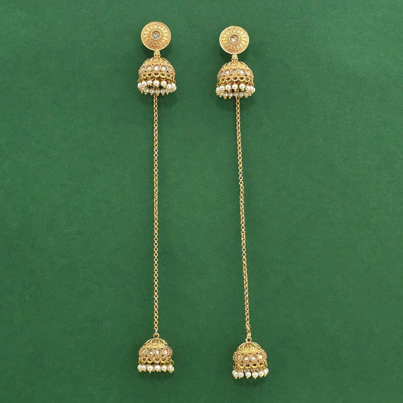 May include: A pair of gold-colored earrings with a floral design and small white pearls. The earrings have a long chain that connects to a bell-shaped charm at the bottom. The background is a solid green color.