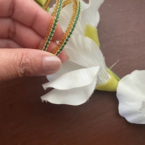 May include: A pair of gold hoop earrings with a green emerald stone trim. The earrings are held in a hand.