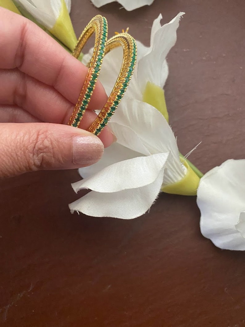 May include: A pair of gold hoop earrings with a green emerald stone trim. The earrings are held in a hand.