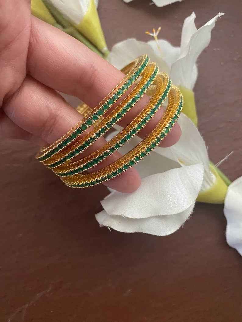 May include: Set of five gold bangles with green gemstone accents. The bangles are stacked on top of each other.