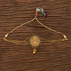 May include: A gold-tone headpiece with a circular design featuring a deity figure surrounded by green and red gemstones. The headpiece has a chain that connects to a dangling tassel with white and red beads.