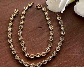 Könnte beinhalten: Ein Paar goldfarbene Fußkettchen mit einer durchgehenden Kette aus ovalen, klaren Steinen in Metallfassungen. Jedes Fußkettchen ist mit einem kleinen S-Haken-Verschluss versehen und zeigt ein wiederkehrendes Muster aus facettierten Edelsteinen.