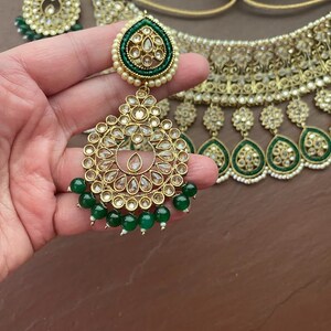 May include: A gold and green Indian bridal necklace set with pearl and rhinestone embellishments. The set includes a choker necklace, earrings, and a maang tikka.