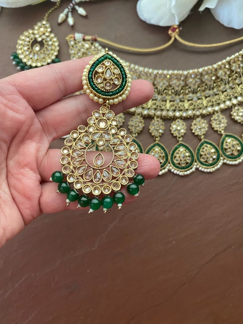 May include: A gold and green Indian bridal necklace set with pearl and rhinestone embellishments. The set includes a choker necklace, earrings, and a maang tikka.