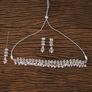 May include: A silver-toned jewelry set featuring a necklace, earrings, and a headpiece. The necklace and earrings have a leaf-like design with small pink beads. The headpiece has a similar design, with a chain and pink beads. The set is displayed on a wooden surface.