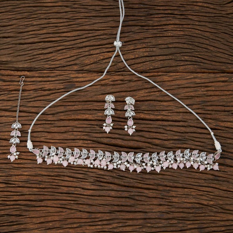 May include: A silver-toned jewelry set featuring a necklace, earrings, and a headpiece. The necklace and earrings have a leaf-like design with small pink beads. The headpiece has a similar design, with a chain and pink beads. The set is displayed on a wooden surface.