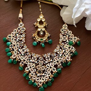 May include: An ornate necklace and headpiece set. The jewelry features a gold-tone metal base with white and black enamel detailing, accented with green gemstone drops. The set is displayed on a brown surface with white flowers in the background.