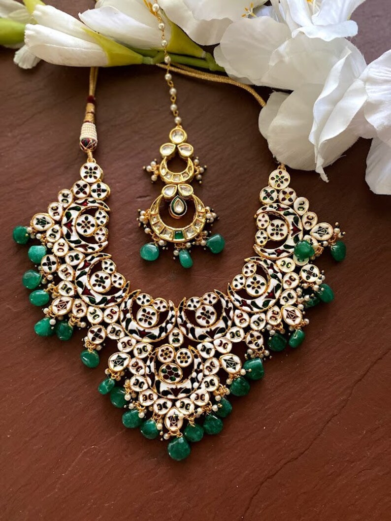 May include: An ornate necklace and headpiece set. The jewelry features a gold-tone metal base with white and black enamel detailing, accented with green gemstone drops. The set is displayed on a brown surface with white flowers in the background.