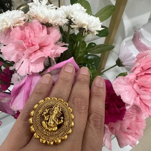 Op de afbeelding: Een grote, ronde goudkleurige ring met een gedetailleerde, in reliëf gemaakte figuur van Ganesha in het midden. De ring heeft een textuurrijke, kralenrand rondom het centrale motief, wat zorgt voor een opvallende, sierlijke uitstraling.