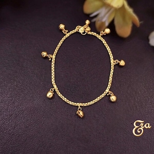 Puede incluir: Una pulsera dorada con un dise&ntilde;o de cadena delicada. La pulsera presenta peque&ntilde;os dijes de campana colgantes. La pulsera se muestra sobre una superficie oscura, con la palabra "Era" en escritura cursiva.