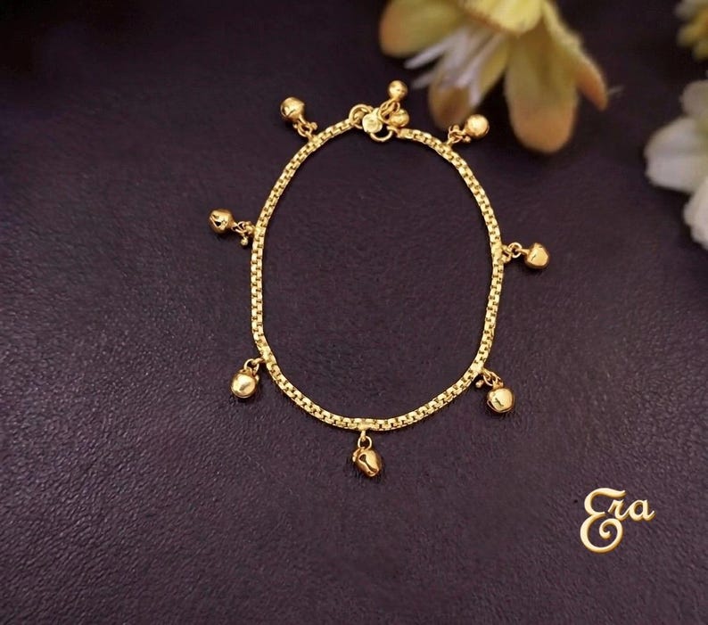 Puede incluir: Una pulsera dorada con un dise&ntilde;o de cadena delicada. La pulsera presenta peque&ntilde;os dijes de campana colgantes. La pulsera se muestra sobre una superficie oscura, con la palabra "Era" en escritura cursiva.