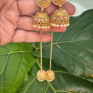 May include: Gold-toned earrings with intricate filigree and small pearl accents. The earrings feature a circular top, a bell-shaped center, and a dangling ball at the bottom. The design is ornate and detailed, with a vintage aesthetic.
