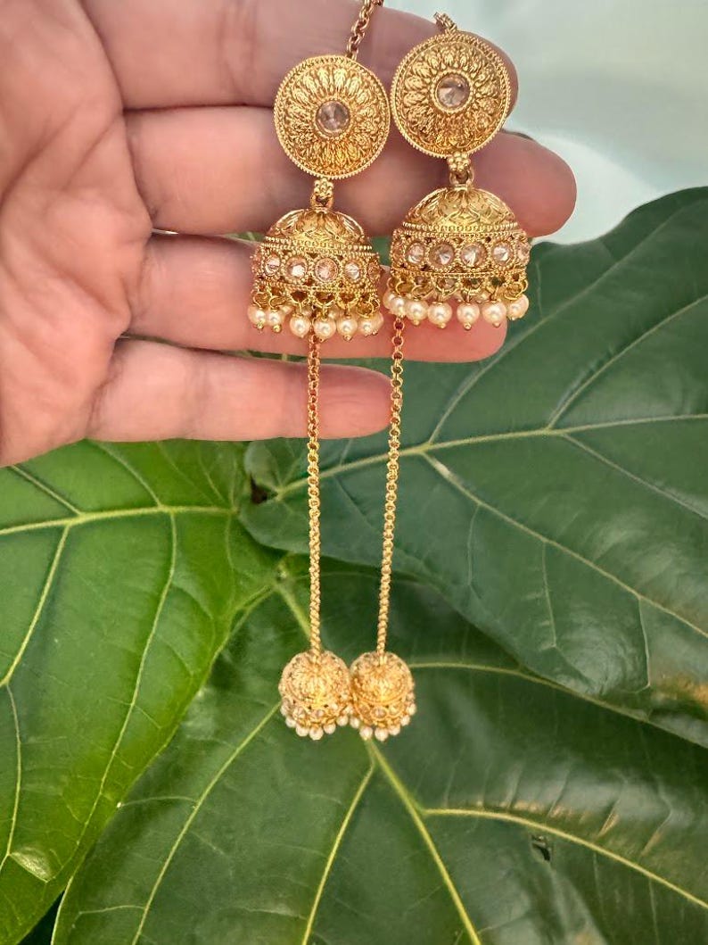 May include: Gold-toned earrings with intricate filigree and small pearl accents. The earrings feature a circular top, a bell-shaped center, and a dangling ball at the bottom. The design is ornate and detailed, with a vintage aesthetic.