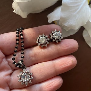 May include: A silver necklace with a round pendant featuring a large clear stone surrounded by smaller stones. The pendant is in the shape of a sun. The necklace is on a black beaded chain. There are also two matching earrings with the same design.