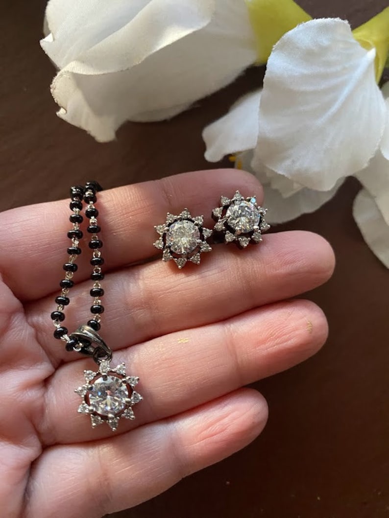 May include: A silver necklace with a round pendant featuring a large clear stone surrounded by smaller stones. The pendant is in the shape of a sun. The necklace is on a black beaded chain. There are also two matching earrings with the same design.