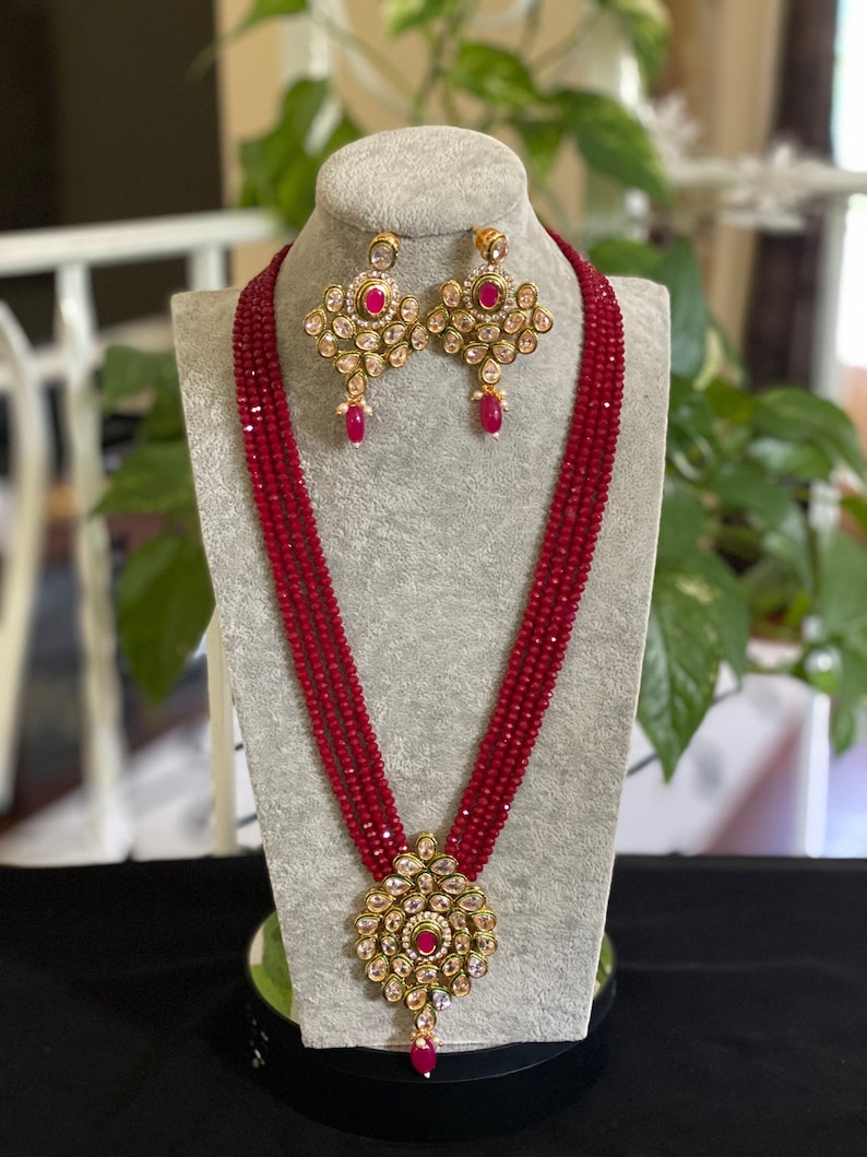 May include: A red beaded necklace with a gold pendant and matching earrings. The pendant and earrings feature a floral design with pink stones.