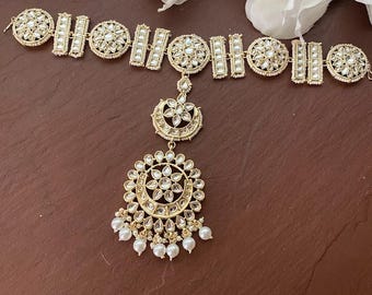 May include: A gold-toned hair accessory featuring a large, round, pearl-encrusted pendant and a smaller, crescent-shaped pendant. The accessory is attached to a chain of smaller, rectangular, pearl-encrusted pieces.