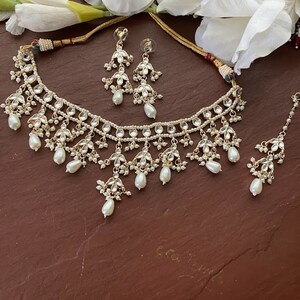 May include: A gold necklace with a multi-layered design featuring white pearls and clear stones. The necklace is paired with matching earrings and a maang tikka.