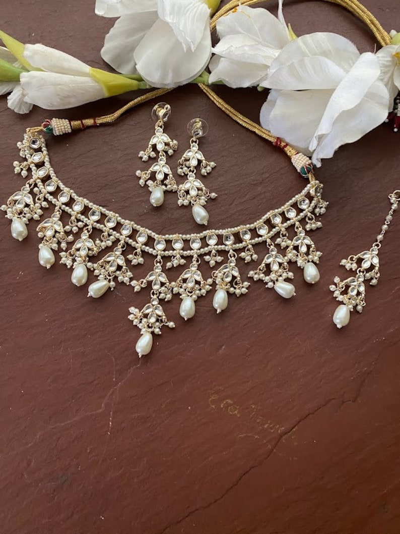 May include: A gold necklace with a multi-layered design featuring white pearls and clear stones. The necklace is paired with matching earrings and a maang tikka.