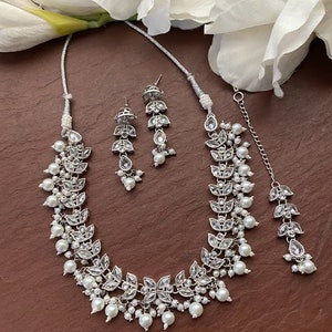 May include: A silver-toned jewelry set featuring a necklace, earrings, and a headpiece. The necklace and headpiece have a leaf design with clear stones and white pearls. The earrings have a similar design. The set is displayed on a brown surface with white flowers.
