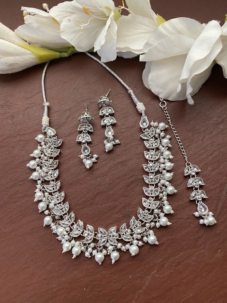 May include: A silver-toned jewelry set featuring a necklace, earrings, and a headpiece. The necklace and headpiece have a leaf design with clear stones and white pearls. The earrings have a similar design. The set is displayed on a brown surface with white flowers.