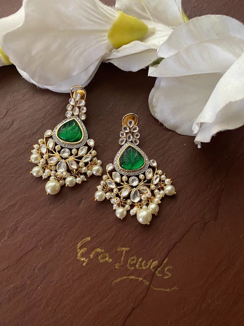 May include: A pair of gold earrings with green gemstone accents and pearl drops. The earrings feature intricate floral designs and are set against a brown background. The text "Era Jewels" is written in cursive on the surface.