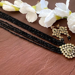 May include: A black beaded necklace with a gold pendant and matching earrings. The pendant is adorned with clear and green stones.