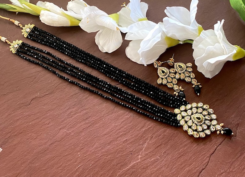 May include: A black beaded necklace with a gold pendant and matching earrings. The pendant is adorned with clear and green stones.