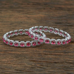 May include: Two silver bangles with pink square-shaped stones set in a silver frame. The bangles are on a wooden surface.