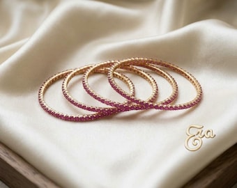 May include: A set of three thin gold bangles, each featuring a continuous row of small, round, vibrant pink gemstones set along the outer surface. The jewellery pieces are arranged in an overlapping circular formation with the text Era visible.