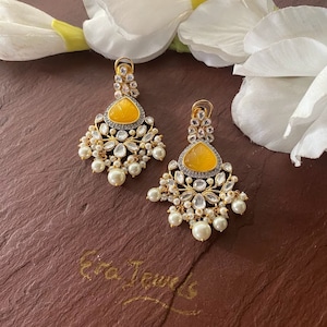 May include: A pair of yellow and white dangle earrings with pearl accents. The earrings feature a yellow stone with a carved design and are surrounded by white stones. The earrings are hanging from a gold-colored metal base.