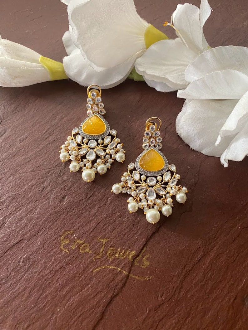 May include: A pair of yellow and white dangle earrings with pearl accents. The earrings feature a yellow stone with a carved design and are surrounded by white stones. The earrings are hanging from a gold-colored metal base.